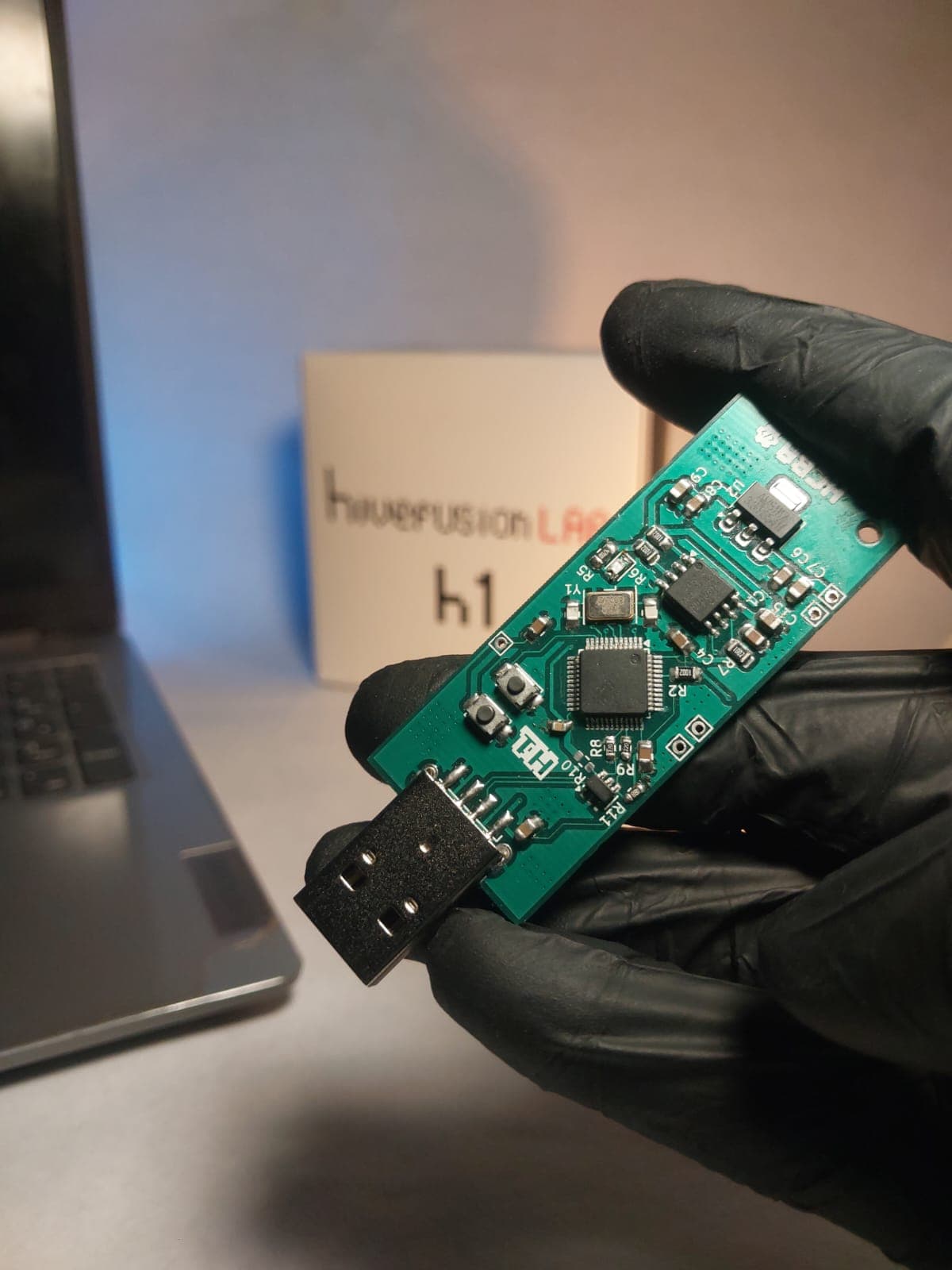The HiveFusion Lab H1 board held in a gloved hand, its USB-A connector facing a laptop.